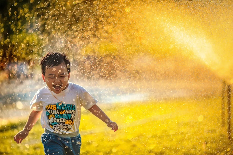 A boy laughing as he runs through a garden sprinkler in golden light